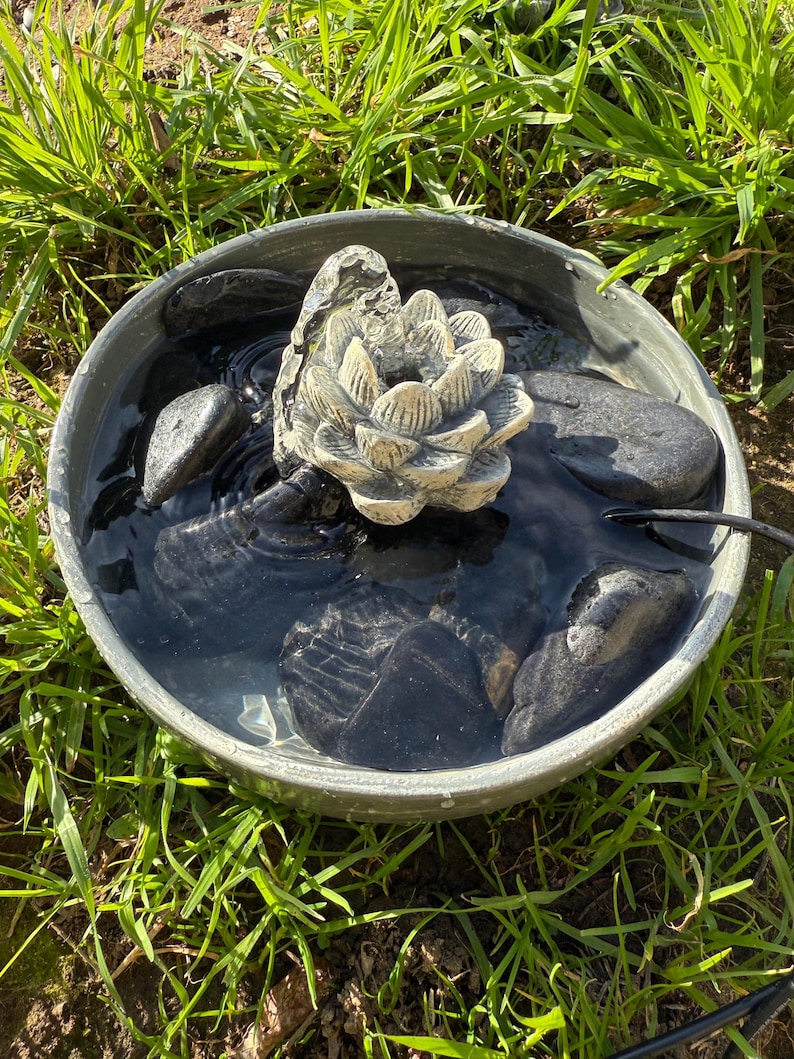 May include: A small, gray, circular water fountain with a white, ceramic lotus flower in the center. The fountain is filled with water and has black stones around the base.