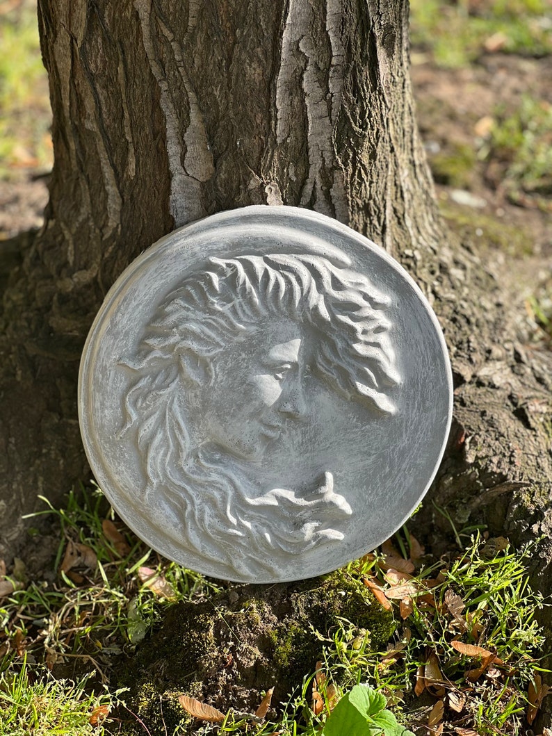 May include: A gray, round, concrete sculpture of a woman's face with flowing hair. The sculpture is mounted on a tree trunk.