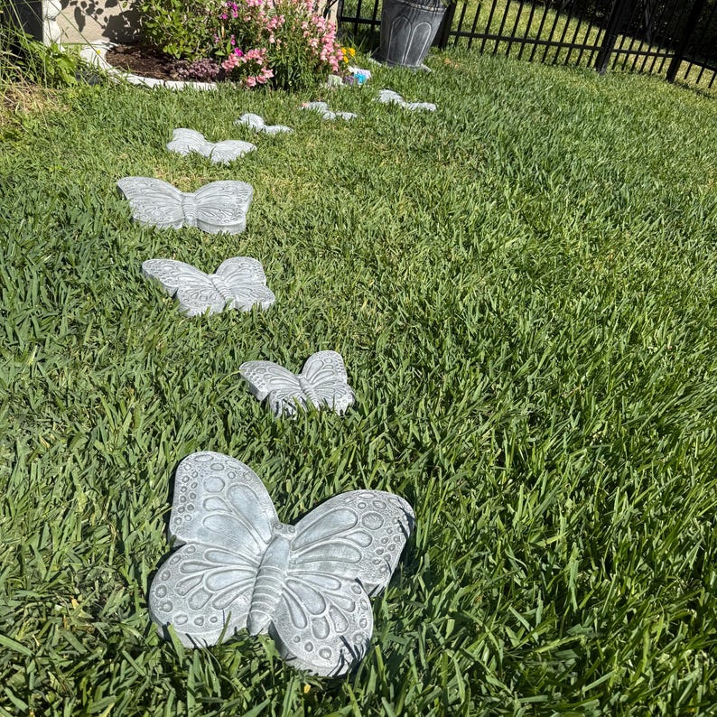 May include: A series of gray butterfly-shaped stepping stones arranged in a grassy yard. The stones vary in size and are made of a textured material. The butterfly garden stones are a decorative element for outdoor spaces.