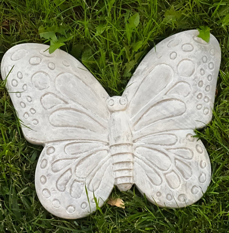 May include: A light gray, concrete butterfly garden ornament. The butterfly has detailed wing patterns and a textured surface. The ornament is resting on a bed of green grass.
