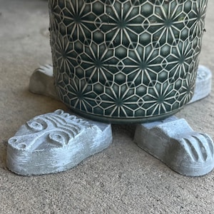 May include: A set of four concrete animal paw-shaped plant pot feet. The feet are painted a light gray color and have a textured surface. A green ceramic pot with a geometric pattern is sitting on top of the feet.