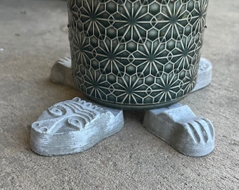 May include: A set of four concrete animal paw-shaped plant pot feet. The feet are painted a light gray color and have a textured surface. A green ceramic pot with a geometric pattern is sitting on top of the feet.