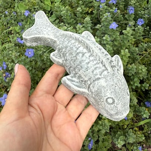 May include: A gray concrete koi fish garden ornament. The fish has detailed fins and scales, and is held in a hand against a backdrop of green foliage and small blue flowers. The fish is approximately 10 inches long.