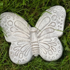 May include: A light gray concrete butterfly garden ornament. The butterfly has detailed wing patterns and is resting on green grass. The wings have a textured surface with circular and curved designs. The butterfly is a decorative garden accent.