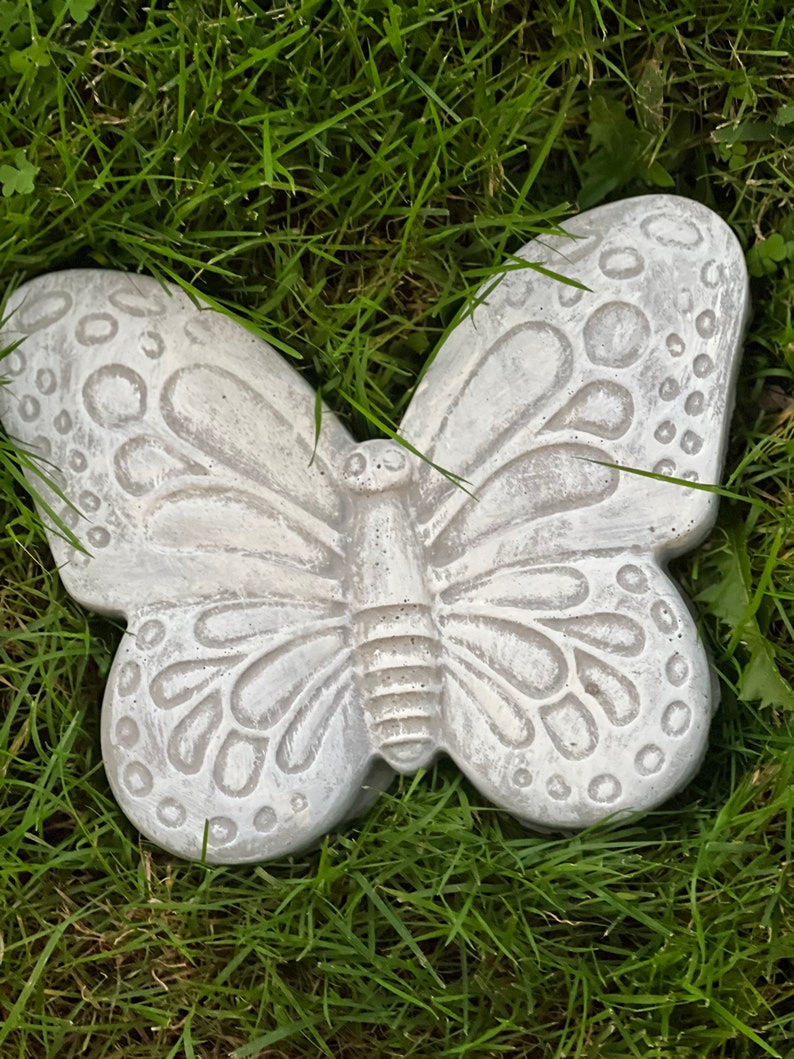 May include: A light gray concrete butterfly garden ornament. The butterfly has detailed wing patterns and is resting on green grass. The wings have a textured surface with circular and curved designs. The butterfly is a decorative garden accent.