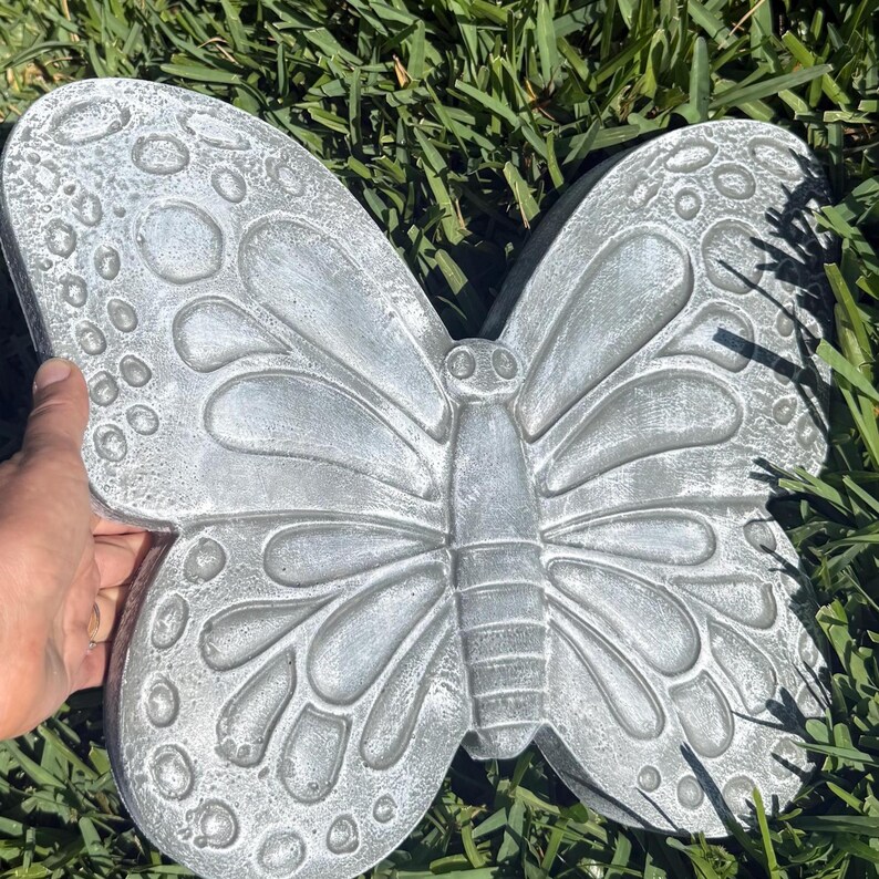 May include: A gray concrete butterfly garden statue with detailed wing patterns and a textured surface. The butterfly is resting on green grass, showcasing its decorative design for outdoor spaces.