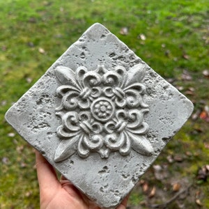 May include: A gray concrete tile with a raised, ornate design in the center. The tile has a rough, textured surface.