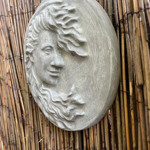 May include: A gray, round, concrete wall sculpture of a woman's face with flowing hair. The sculpture is mounted on a woven bamboo wall.