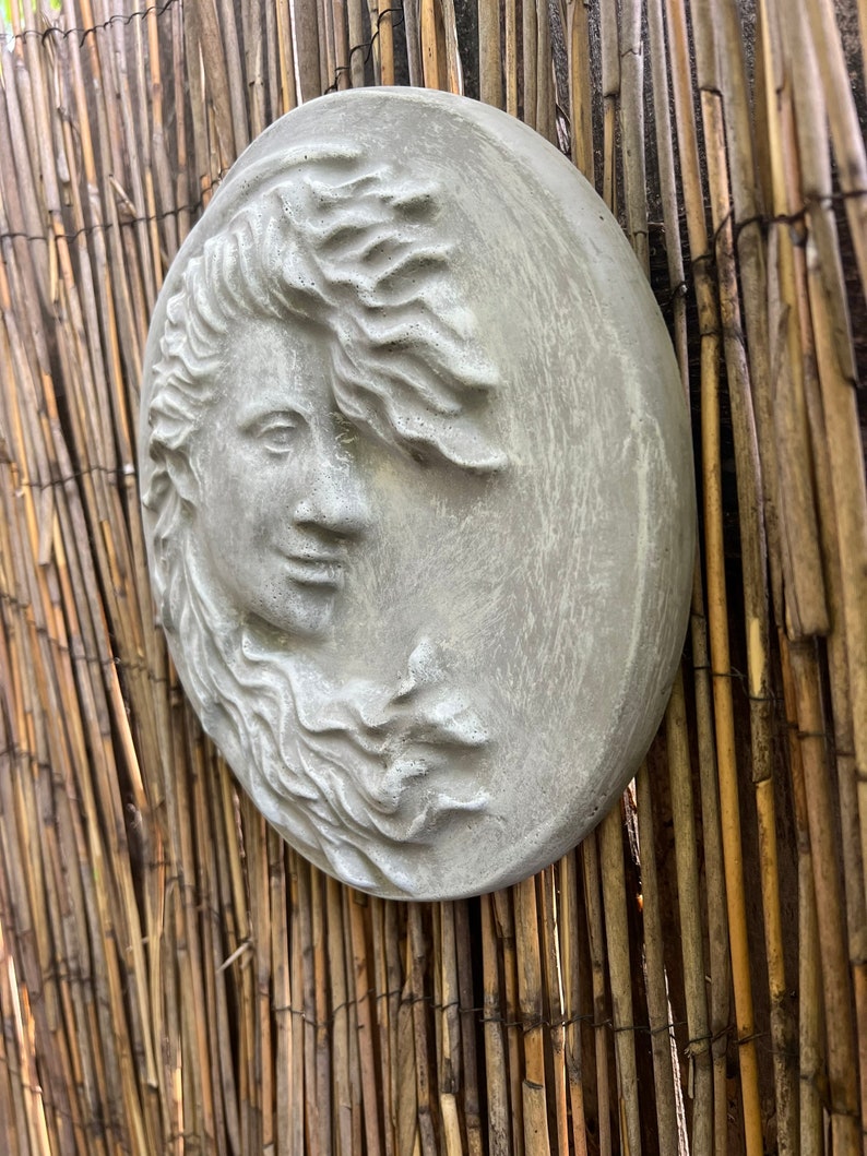 May include: A gray, round, concrete wall sculpture of a woman's face with flowing hair. The sculpture is mounted on a woven bamboo wall.