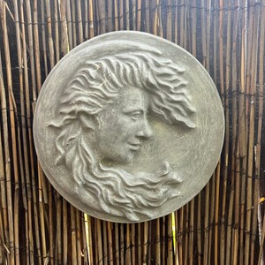May include: A gray stone relief sculpture of a woman's face with flowing hair. The sculpture is circular and has a textured surface.
