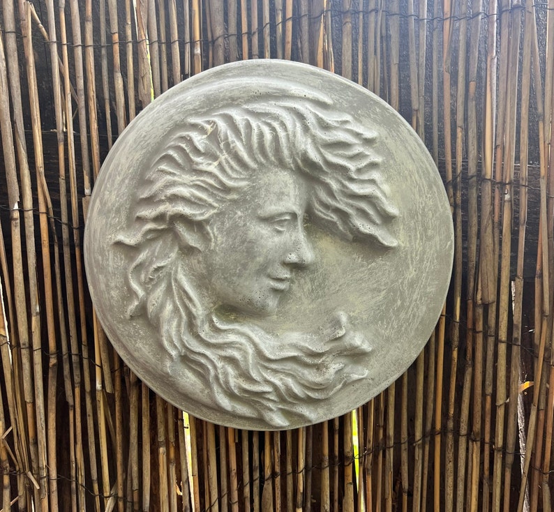 May include: A gray stone relief sculpture of a woman's face with flowing hair. The sculpture is circular and has a textured surface.