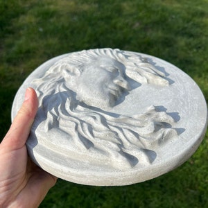 May include: A round, gray concrete wall plaque with a raised, detailed sculpture of a woman's face with flowing hair.