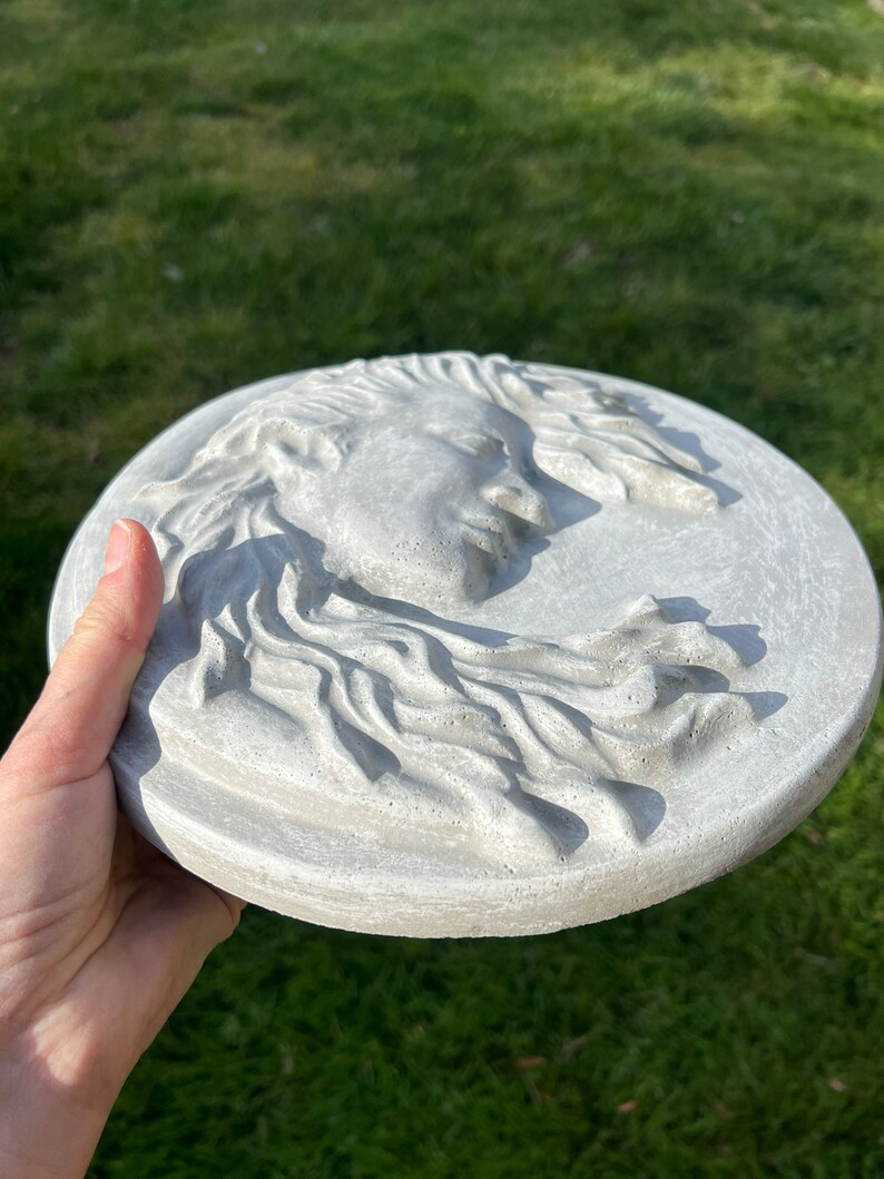 May include: A round, gray concrete wall plaque with a raised, detailed sculpture of a woman's face with flowing hair.