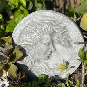 May include: A round, gray stone sculpture of a woman's face with flowing hair. The sculpture is set against a background of green plants.