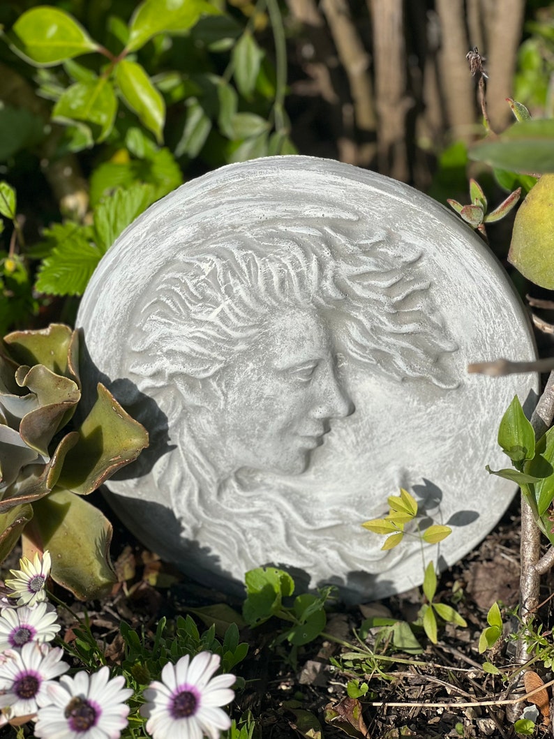 May include: A round, gray stone sculpture of a woman's face with flowing hair. The sculpture is set against a background of green plants.