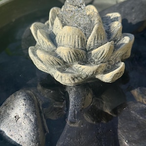 May include: A small, gray, stone fountain with a lotus flower design. Water flows from the top of the flower, creating a relaxing sound. The fountain is surrounded by black stones.