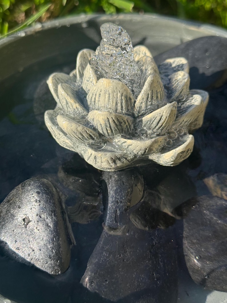 May include: A small, gray, stone fountain with a lotus flower design. Water flows from the top of the flower, creating a relaxing sound. The fountain is surrounded by black stones.