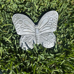 May include: A detailed, silver-colored butterfly garden ornament. The butterfly has intricate wing patterns and is resting on a bed of green grass. The ornament is made of a durable material and is designed for outdoor use.