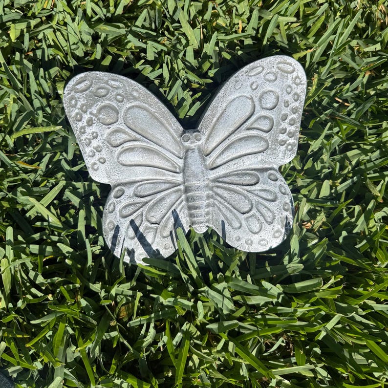 May include: A detailed, silver-colored butterfly garden ornament. The butterfly has intricate wing patterns and is resting on a bed of green grass. The ornament is made of a durable material and is designed for outdoor use.