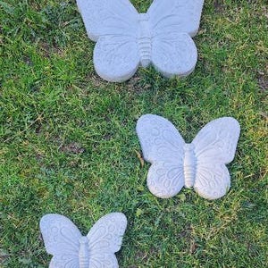 May include: Three gray butterfly garden ornaments of varying sizes rest on green grass. Each butterfly has detailed wing patterns and a textured surface. The butterflies are arranged in a vertical composition.