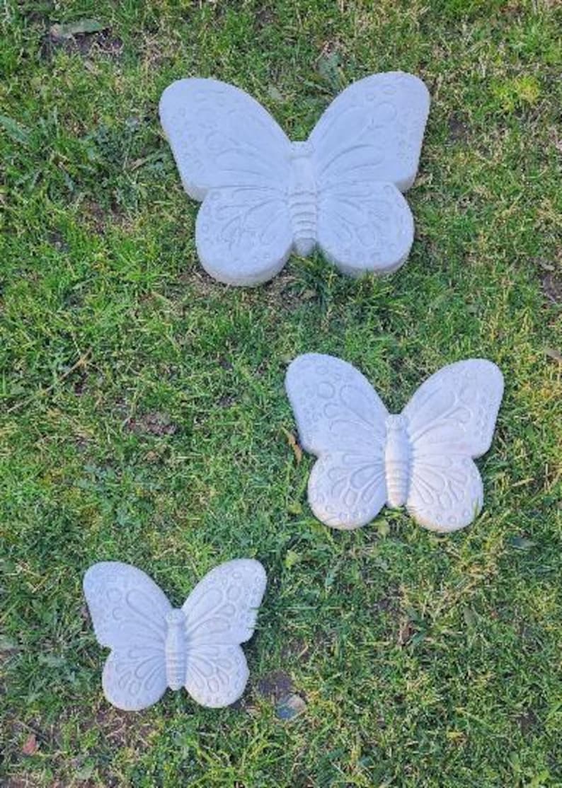 May include: Three gray butterfly garden ornaments of varying sizes rest on green grass. Each butterfly has detailed wing patterns and a textured surface. The butterflies are arranged in a vertical composition.