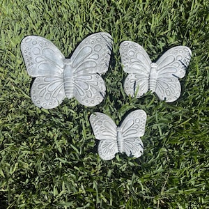 May include: Three gray butterfly garden ornaments of varying sizes are arranged on green grass. The butterflies have detailed wing patterns and are made of a solid material. These decorative pieces are suitable for outdoor use.