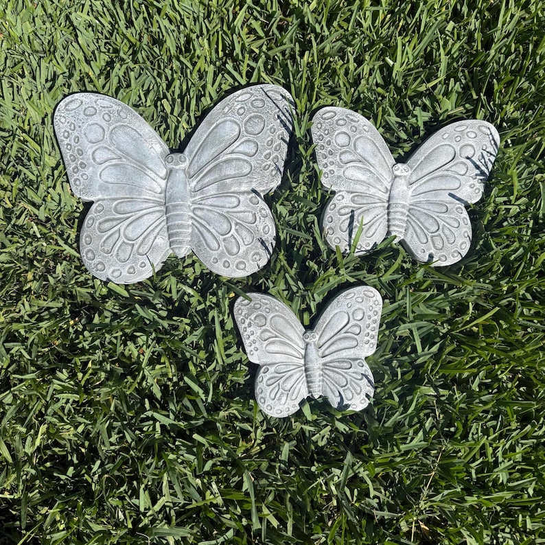May include: Three gray butterfly garden ornaments of varying sizes are arranged on green grass. The butterflies have detailed wing patterns and are made of a solid material. These decorative pieces are suitable for outdoor use.