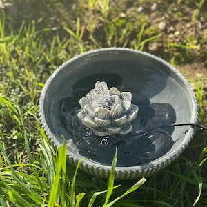May include: A gray ceramic bird bath with a water fountain featuring a lotus flower design. The fountain is surrounded by green grass.