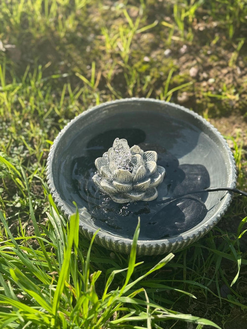 May include: A gray ceramic bird bath with a water fountain featuring a lotus flower design. The fountain is surrounded by green grass.