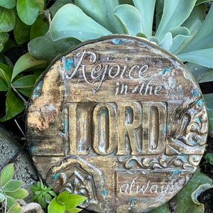 May include: A round, rustic, gold-colored stone with the words "Rejoice in the LORD always" carved into it. The stone has a distressed, aged look with a teal-colored paint accent.