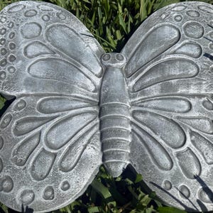 May include: A gray concrete butterfly garden ornament. The butterfly has detailed wing patterns and a textured surface. The wings are spread wide, and the body is segmented. The ornament is designed to be placed outdoors.