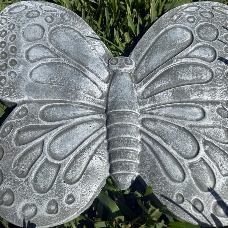 May include: A gray concrete butterfly garden ornament. The butterfly has detailed wing patterns and a textured surface. The wings are spread wide, and the body is segmented. The ornament is designed to be placed outdoors.