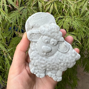 Puede incluir: Una figurita de oveja de hormig&oacute;n gris con una superficie texturizada. La oveja tiene una cabeza redonda con una nariz peque&ntilde;a y ojos grandes. La oveja est&aacute; de pie sobre cuatro patas.