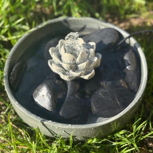 May include: A small, gray, circular fountain with a white, flower-shaped spout. The fountain is filled with water and has black stones around the base. The fountain is sitting on a bed of green grass.