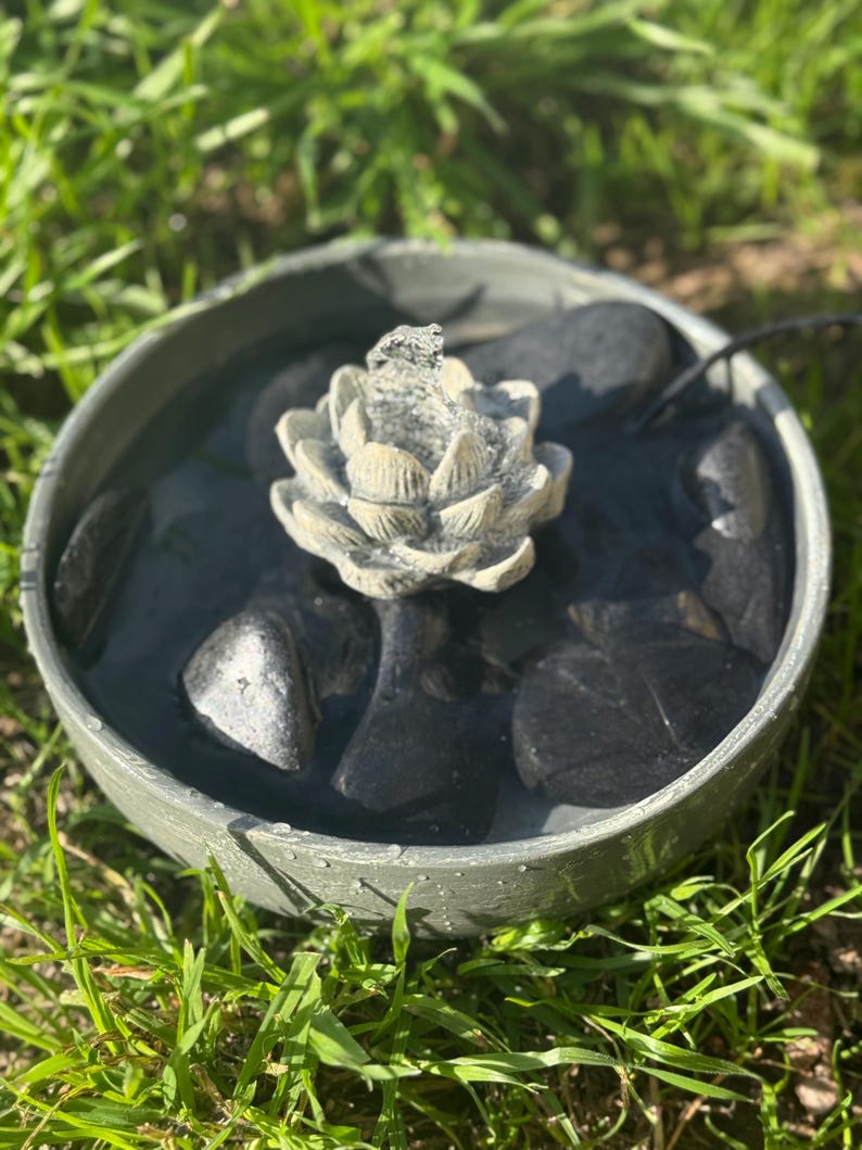 May include: A small, gray, circular fountain with a white, flower-shaped spout. The fountain is filled with water and has black stones around the base. The fountain is sitting on a bed of green grass.