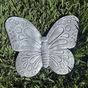 May include: A gray butterfly-shaped garden ornament. The butterfly has detailed wing patterns with circular and curved designs. The ornament is made of a solid material and is resting on green grass.