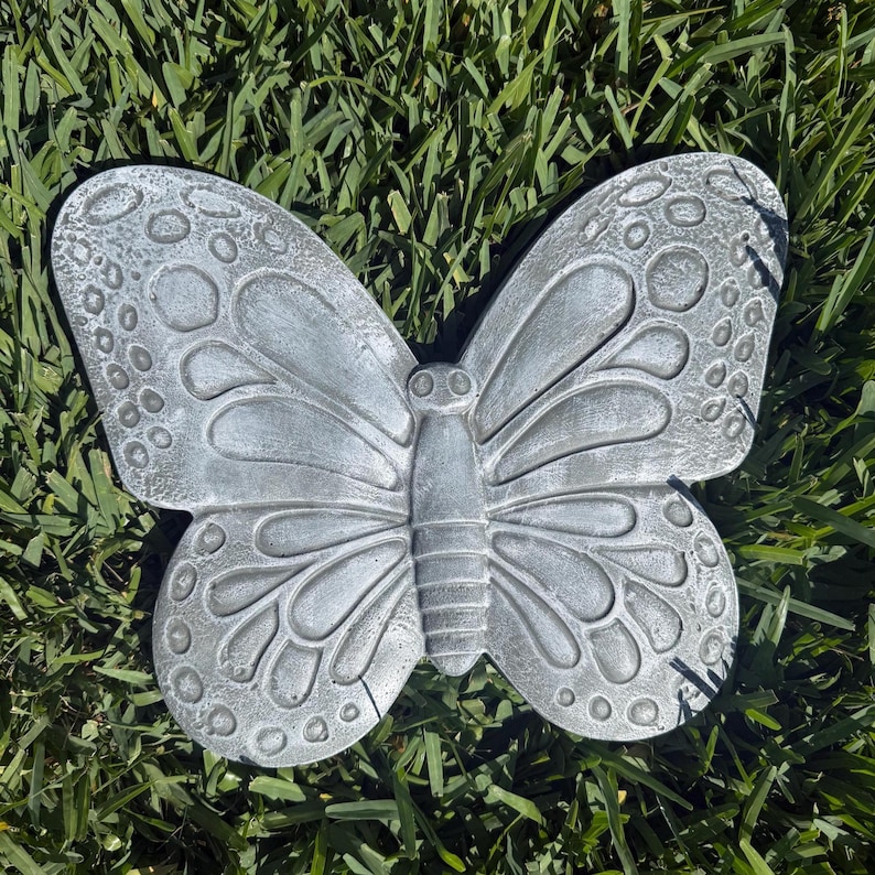 May include: A gray butterfly-shaped garden ornament. The butterfly has detailed wing patterns with circular and curved designs. The ornament is made of a solid material and is resting on green grass.