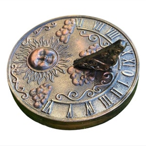May include: A bronze-colored sundial with Roman numerals and a decorative design featuring grapes and a sun. The sundial has a copper-colored gnomon.