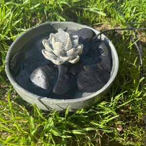 May include: A small gray concrete fountain with a white ceramic lotus flower in the center. The fountain is filled with water and has black stones around the base.