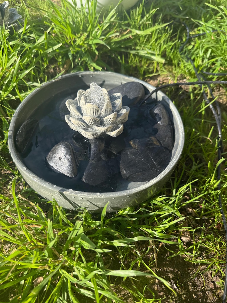 May include: A small gray concrete fountain with a white ceramic lotus flower in the center. The fountain is filled with water and has black stones around the base.