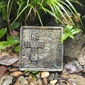 May include: A gold and blue metal plaque with a cross and the text "Life is Fragile handle with Prayer". The plaque is surrounded by small rocks and green plants.