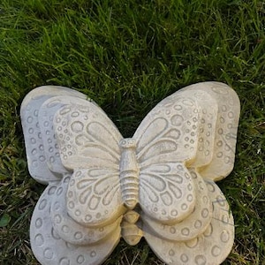 May include: A light gray, stone-like butterfly sculpture with detailed wing patterns and a textured surface. The butterfly is resting on a bed of green grass, showcasing its decorative design. This garden decor piece adds a touch of nature to any outdoor space.