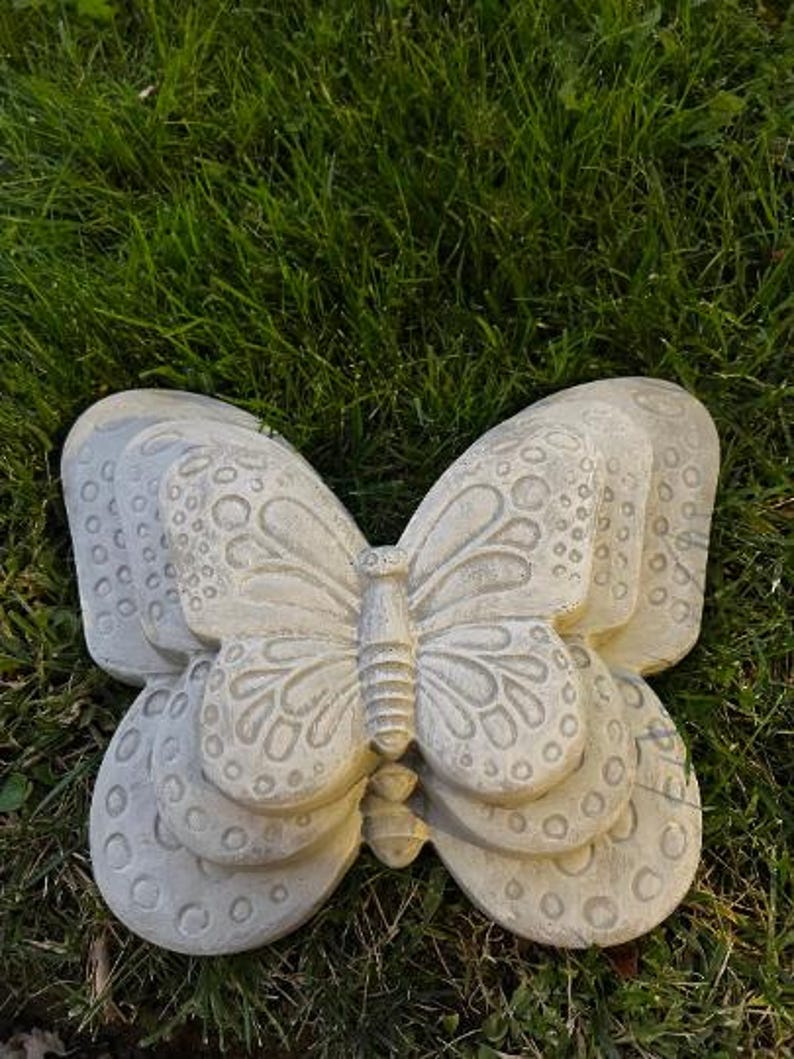 May include: A light gray, stone-like butterfly sculpture with detailed wing patterns and a textured surface. The butterfly is resting on a bed of green grass, showcasing its decorative design. This garden decor piece adds a touch of nature to any outdoor space.