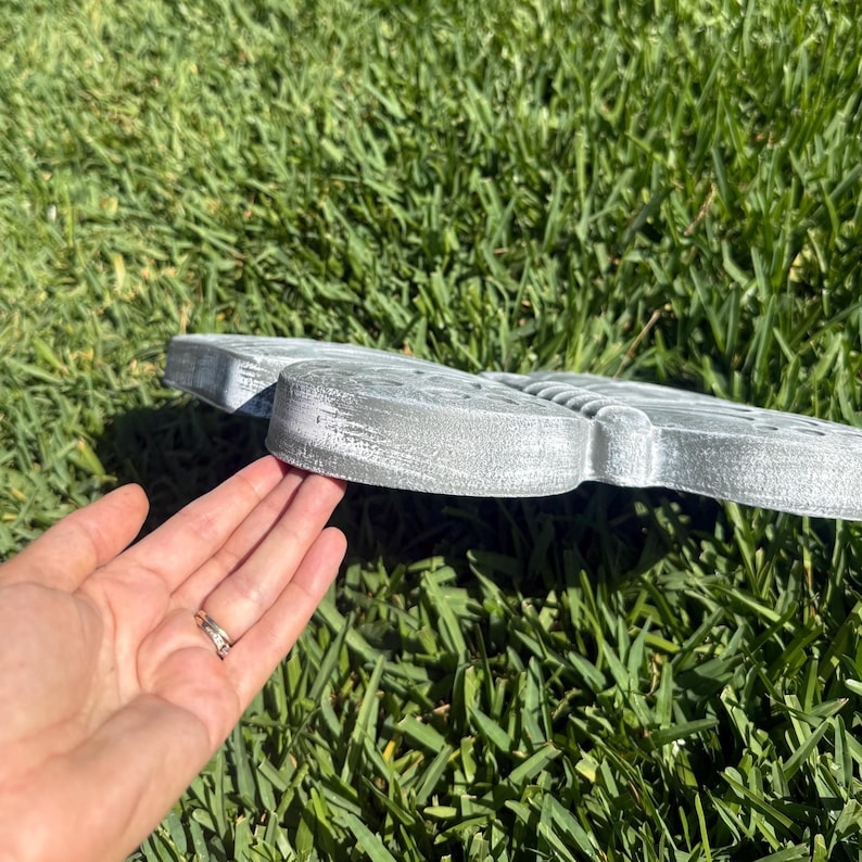 May include: A gray, butterfly-shaped garden stepping stone. The stone has a textured surface and is resting on green grass. The butterfly design is visible on the top surface of the stone.