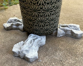 May include: Three concrete plant stands in the shape of a foot, supporting a black and white patterned ceramic pot.