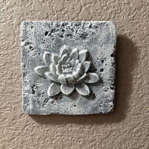 May include: A square, gray concrete wall plaque with a raised, white, sculpted flower design. The flower has a central circle with five petals.