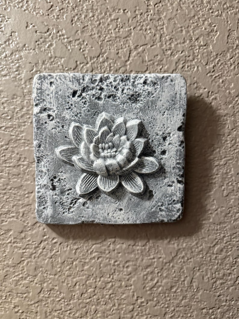 May include: A square, gray concrete wall plaque with a raised, white, sculpted flower design. The flower has a central circle with five petals.