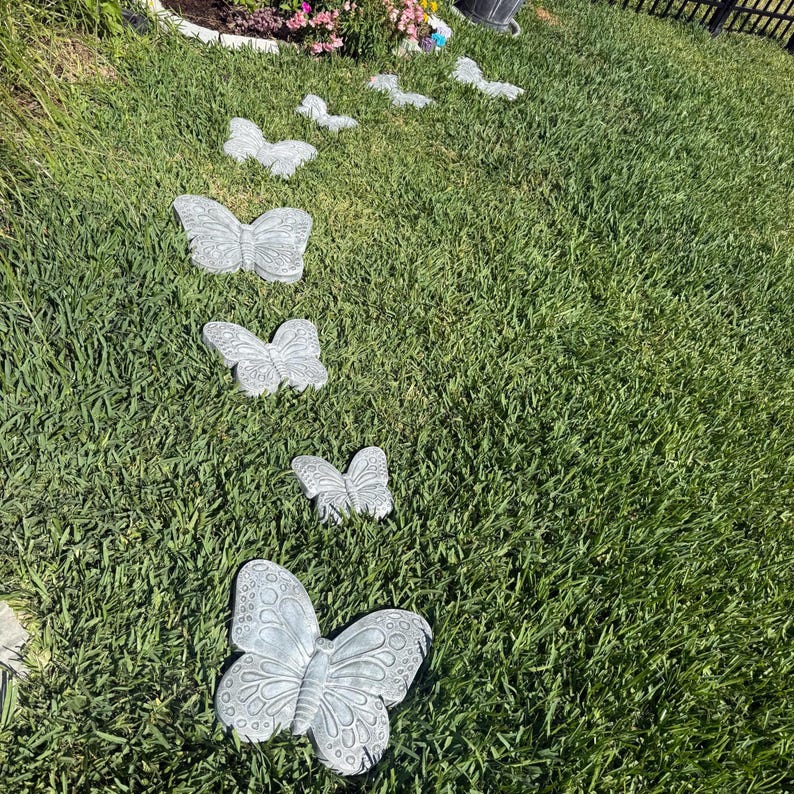 May include: A series of gray butterfly-shaped stepping stones arranged in a line across a green lawn. The stones vary in size, with detailed wing patterns. The scene is outdoors, with grass and a flower bed visible.