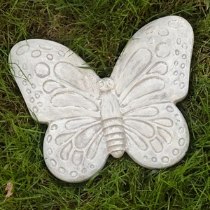 May include: A detailed, off-white butterfly garden ornament. The butterfly has textured wings with circular patterns and detailed vein lines. The sculpture is resting on a bed of green grass.