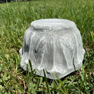 May include: A gray, textured concrete planter, shaped like a small, upside-down bowl. The planter sits in green grass, showcasing its rough, weathered surface. The design is simple and rustic, suitable for small plants or succulents.